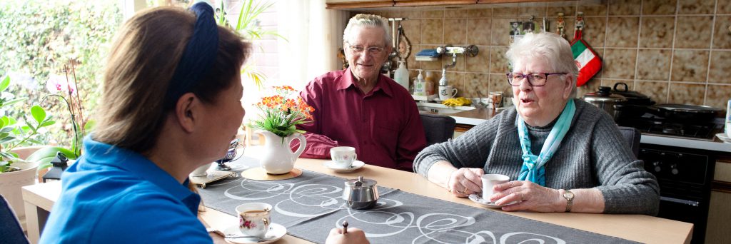 Huishoudelijke hulp ondersteunt oudere echtpaar tijdens koffietijd in eigen keuken thuis